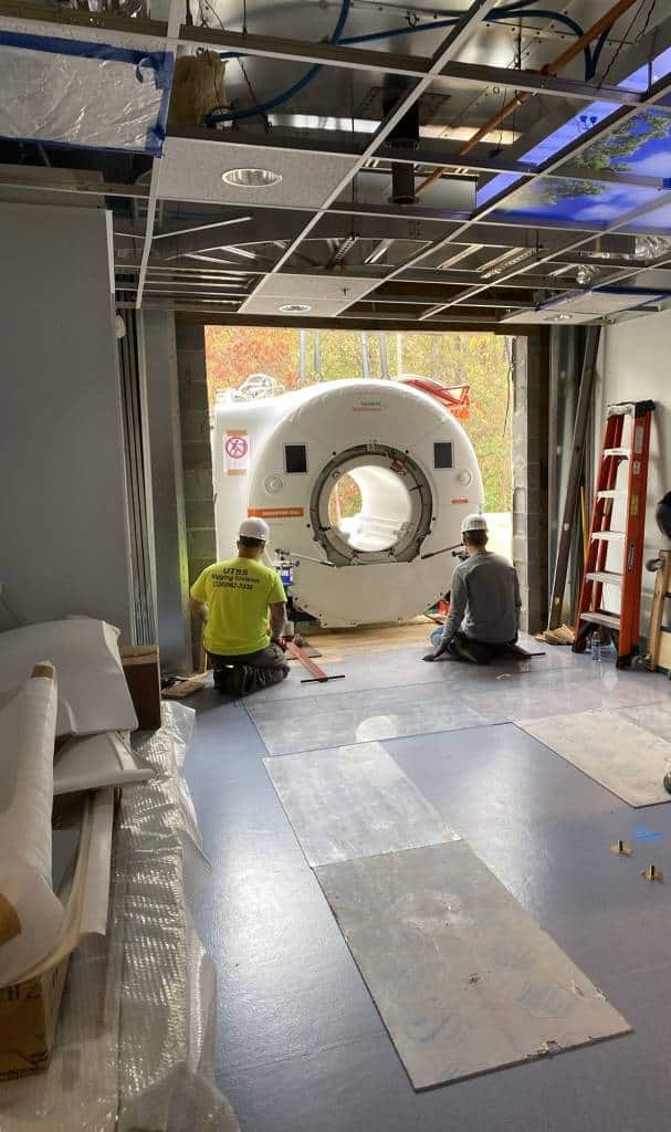 two construction men sitting on concrete floor waiting to move an MRI machine into a large opening into a building.