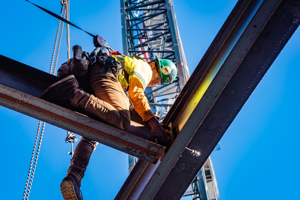A worker in a hard hat and harness installs a steel beam while standing on another beam