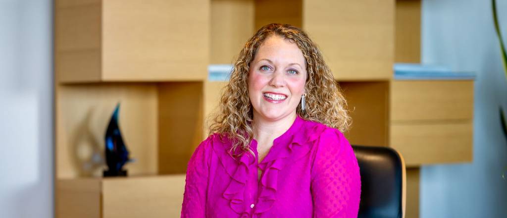 Women with blonde curly hair, wearing a pink blouse and blue blazer smiles straight ahead white sitting in a black leather chair.