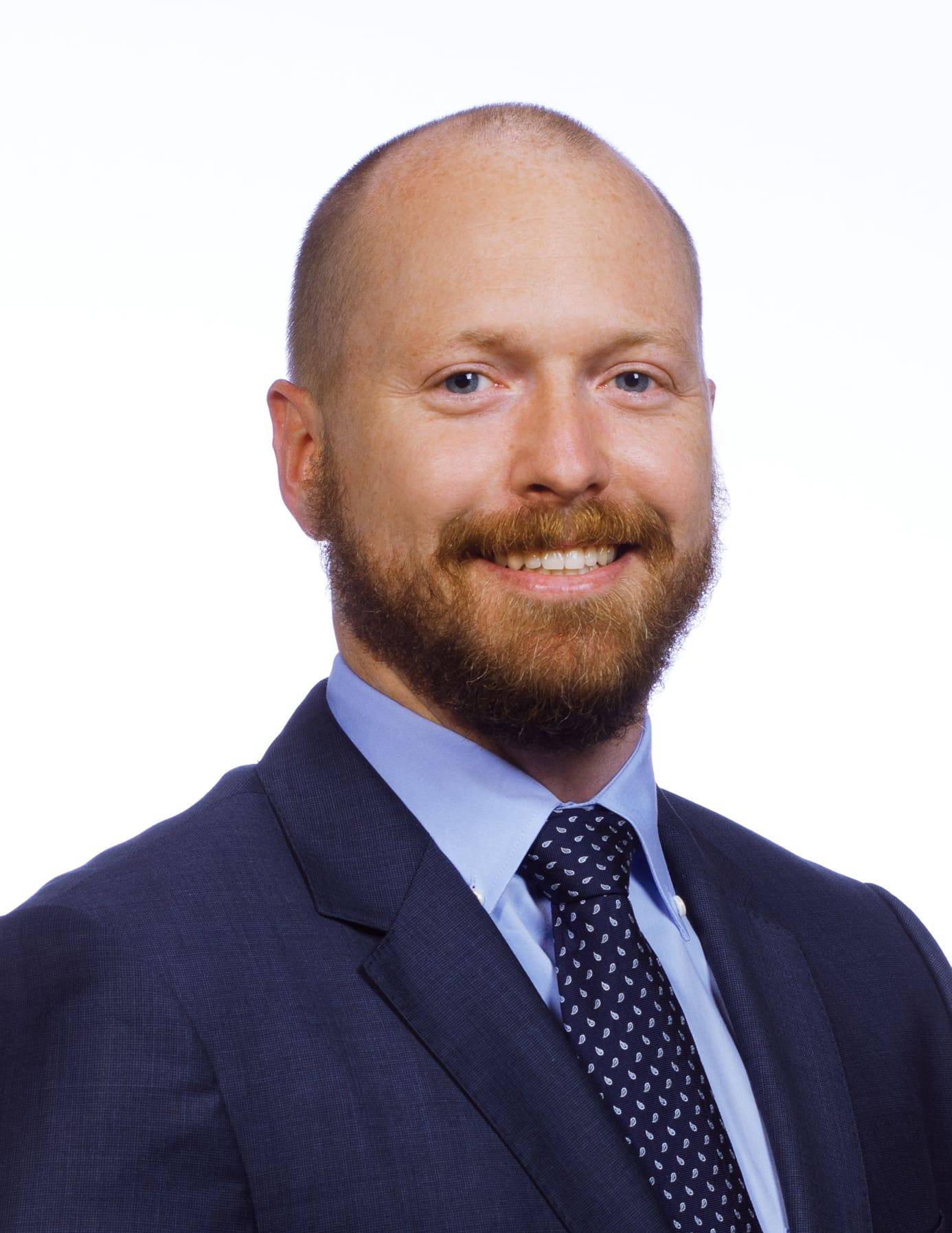 Man with bead wearing a suit and tie smiles.